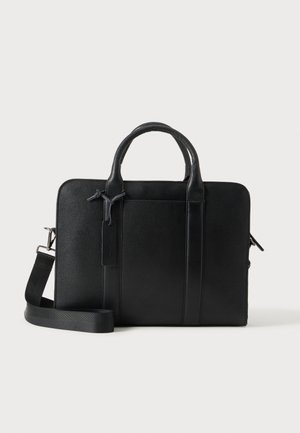 Mallette en cuir noir avec doubles poignées, bandoulière détachable, poche avant et fermeture éclair sur fond clair.