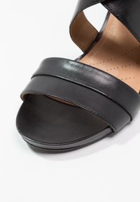 Sorte lædersandaler med bred stropdesign, åben tå og glat tekstur. Lys tan indersål med perforeringsdetaljer for åndbarhed.