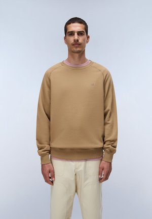 Mann, der nach vorn schaut, trägt einen hellbraunen Pullover und cremefarbene Hose vor einem schlichten hellen Hintergrund.