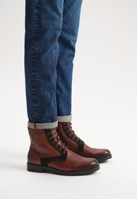 Brązowe skórzane botki za kostkę z wiązaniem i zamszowymi akcentami. Gładka tekstura; okrągły nosek; noszone z podwiniętymi dżinsami.