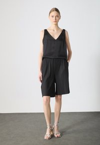 Black sleeveless top with a V-neck and two side pockets, paired with pleated black shorts. Model wears silver strappy sandals.