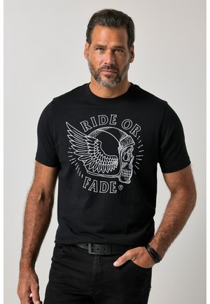 Uomo che indossa una maglietta nera con una grafica bianca di casco con teschio alato e testo "Ride or Fade", in piedi con una mano in tasca.