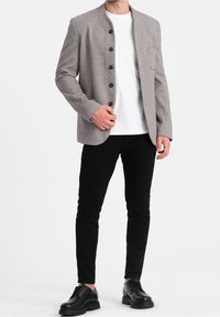 Giacca grigia a bottoni, con un motivo a spina di pesce, caratterizzata da un colletto alla cinese e bottoni neri, indossata sopra una maglietta bianca e pantaloni neri.