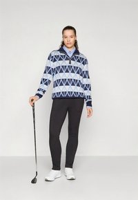 Blå och vitmönstrad zip-up tröja med geometrisk design, kombinerad med svarta leggings och vita skor. Modellen håller i en golfklubba.