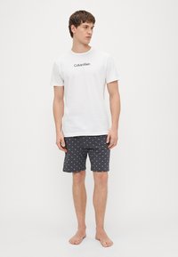 Bela bombažna majica s črnim logotipom "Calvin Klein", kombinirana s temnosivimi kopalkami z belim zvezdnim vzorcem.