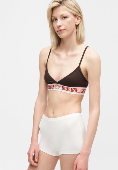 Ξανθιά γυναίκα που φοράει καφέ μπραλέτα με κόκκινη ζώνη με σχέδια και λευκά σορτς ψηλόμεσα, στέκεται μπροστά σε απλό γκρι φόντο.