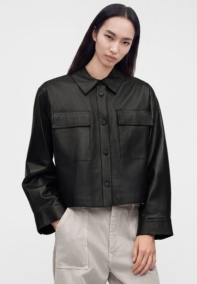 Woman with long dark hair wearing a buttoned black leather jacket with two front pockets and light beige pants with pockets.