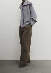 Chemise à rayures avec un grand col, associée à un pantalon large marron et des chaussures noires, le tout sur un fond neutre.