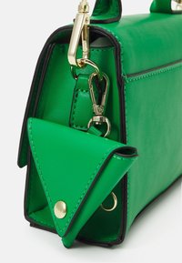 Vihreä nahkakäsilaukku mustalla reunuksella ja kolmionmuotoinen vihreä kolikoiden pussi, joka on kiinnitetty kultaklipsuilla ja napin painikkeella.