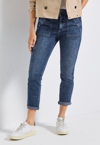 Dunkelblaue, leicht distressed Denim-Jeans mit aufgerollten Säumen, ausgestattet mit Fronttaschen und einem figurbetonten Schnitt. Kombiniert mit weißen Sneakers.