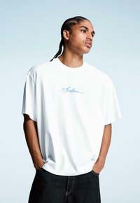 Tricou alb din bumbac, cu croială lejeră, decolteu rotund și mâneci scurte; are text brodat de culoare albastră "Value" pe piept, asortat cu blugi negri.