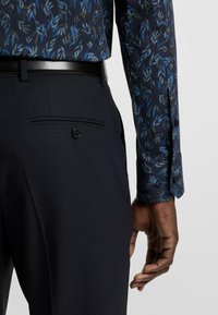 Marineblauwe bloemenoverhemd met blauwe en groene bladmotieven. Donkere broek van fijn geweven stof met een achterzak en een zwarte knop.