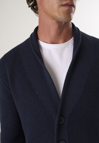 Tumes sweater, mille tekstuuriga, šaalikaeluse ja kolme nupuga. Kantakse valge T-särgi peal, rõhutades selle kihilist disaini.