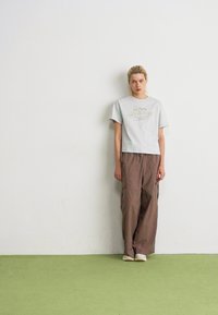 Jeune personne aux cheveux blonds se tient contre un mur blanc, portant un t-shirt gris clair de New Balance, un pantalon brun ample et des chaussures blanches sur un sol vert.