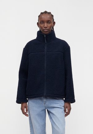 Personne aux cheveux tressés portant une veste polaire bleu marine foncé avec un col montant et un jean bleu clair, debout devant un fond uni.