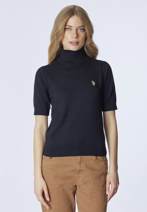 Maglione a collo alto a maniche corte di colore blu marino in maglia liscia; presenta un logo ricamato in oro sul petto. Abbinato a pantaloni marrone chiaro.