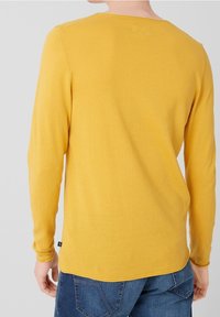 Personne portant un pull jaune moutarde à manches longues et un jean bleu, vue de dos sur un fond clair uni.