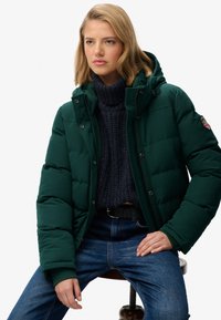 Zielona puchowa kurtka z kapturem, zapinana na zamek błyskawiczny z guzikami jako ozdobą. Nosi się ją na ciemnym dzianinowym swetrze i niebieskich dżinsach.