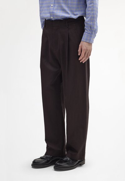 Sötétbarna pliszírozott nadrág puha anyagból, egyenes szárú kialakítással és oldalzsebekkel, fekete bőrcipővel párosítva.