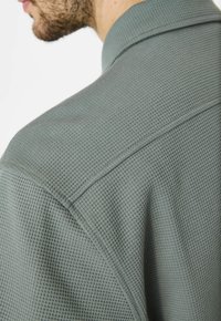 Camisa gris texturizada con un patrón de cuadros, con cuello y detalles de costura visibles en el hombro y la costura trasera.