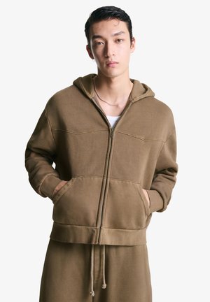 Bruine rits-hoodie met een ontspannen pasvorm, koord onderaan, voorzakken en een textielstructuur. Bevat een gevoerde kap en stikselsaccenten.