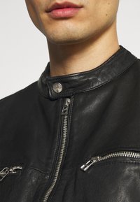 Černá kožená bunda s vysokým límcem, stříbrnými kovovými doplňky a několika kapsami na zip vpředu. Hladká textura s detailním prošíváním.