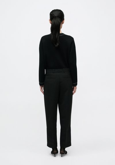 Fekete hosszú ujjú pulóver, fekete magasderekú nadrág és nyitott orrú magas sarkú cipő, hátulnézetből, egyszínű fehér háttér előtt.