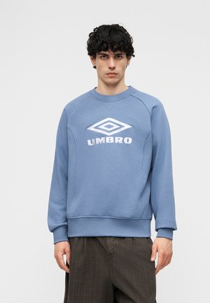 Młody mężczyzna z kręconymi włosami, ubrany w niebieską bluzę Umbro i ciemne spodnie, stojący na tle jednolitego, jasnego tła.