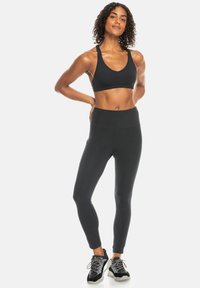 Schwarzer Sport-BH mit breiten Trägern, kombiniert mit hoch taillierten schwarzen Leggings. Texturierte Sneakers mit schwarzen und grauen Akzenten runden den Look ab.