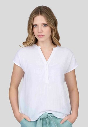 Blouse blanche à manches courtes avec un col en V et des boutons, réalisée dans un tissu texturé subtil. Le mannequin porte un pantalon vert clair, les mains dans les poches.