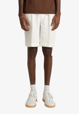 Uomo che indossa pantaloncini bianchi fino al ginocchio, calzini bianchi corti e sneakers bianche con suola in gomma, in piedi su uno sfondo bianco semplice.