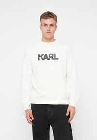 Mladý muž v bielom polo-krku s logom "KARL LAGERFELD" a čiernymi nohavicami, stojaci na jednofarebnom bielom pozadí.