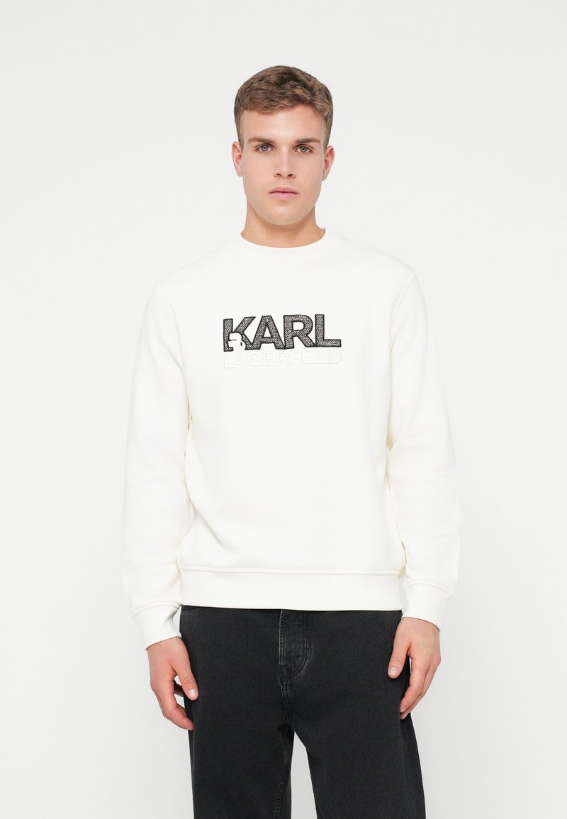 Mladý muž v bielom polo-krku s logom "KARL LAGERFELD" a čiernymi nohavicami, stojaci na jednofarebnom bielom pozadí.