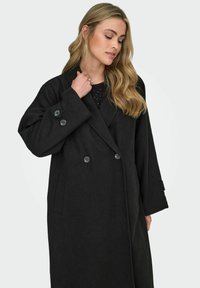 Juodas vilnos mišinio paltas su dvigubo sutvarkymo dizainu, dideliais juodais mygtukais ir šoninėmis kišenėmis. Turi pritaikytą apykaklę ir ilgus rankovės.