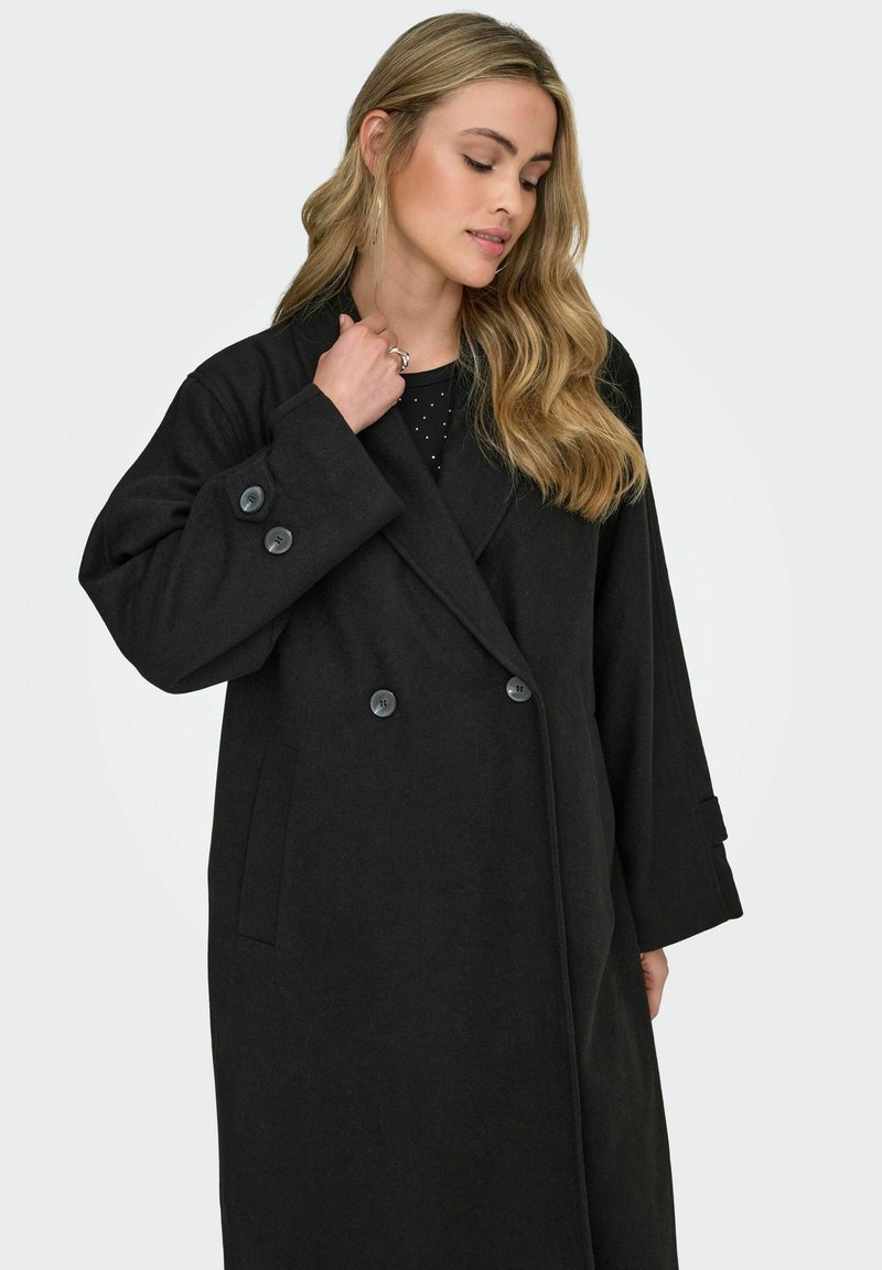 Juodas vilnos mišinio paltas su dvigubo sutvarkymo dizainu, dideliais juodais mygtukais ir šoninėmis kišenėmis. Turi pritaikytą apykaklę ir ilgus rankovės.