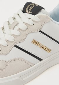 Fehér bőrből és szürke velúrból készült sneaker, fekete csíkokkal, arany logóval és lapos fehér fűzőkkel. A textúrázott talp látható.