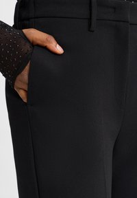 Zwarte broek met een gladde stofafwerking, voorzien van een zijzak en een hand die erin rust. De zoom heeft een doorschijnende, gestippelde textuur.