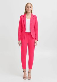 Fitted roze blazer en bijpassende joggers van soepel materiaal. De blazer heeft een enkele knoop, en de joggers hebben een tailleband met trekkoord.