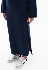 Lang, marineblauwe jurk met een zijsplit en lange mouwen, gemaakt van zachte stof. Draag met witte en grijze sneakers met zwarte accenten.