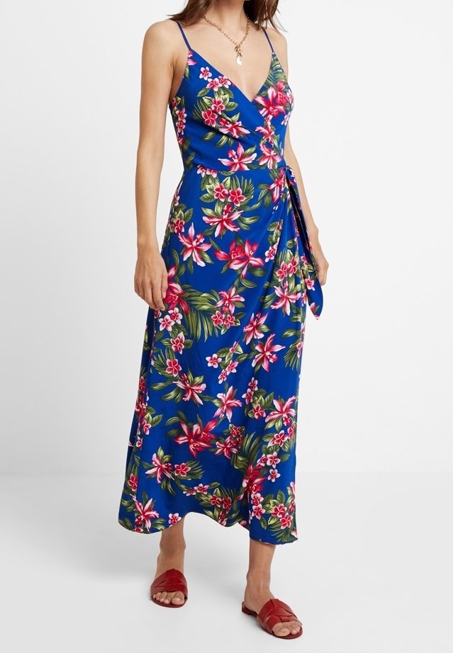 Femme portant une robe portefeuille bleue à fleurs roses et vertes, fines bretelles, et des sandales rouges, debout devant un fond uni.