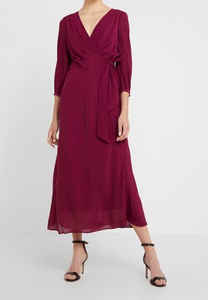 Mujer que lleva un vestido cruzado de manga larga, rojo intenso, con un lazo lateral y sandalias negras de tacón alto, de pie sobre un fondo claro.
