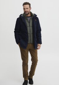 Navy hooded jacket over a green padded vest, beige corduroy pants, and black boots. Features zippers and pockets, with a textured finish.
