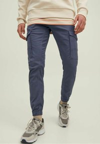 Muž v modrých cargo jogger kalhotách, béžových a šedých teniskách a krémové mikině se pruhovaným lemem, stojící na jednobarevném pozadí.