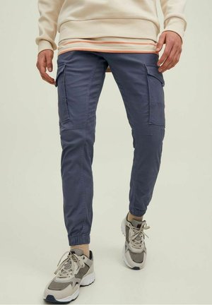 Mężczyzna w niebieskich joggerach cargo, beżowych i szarych sneakersach oraz kremowym swetrze z prążkowanym wykończeniem, stojący na tle jednolitego tła.