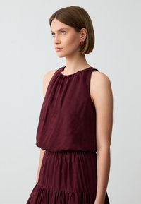 Top senza maniche bordeaux con scollatura arricciata e vestibilità ampia, caratterizzato da un design a gonna a strati e dettagli con bottoni lungo il lato.