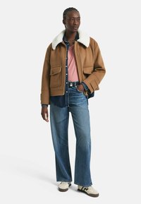 Beige wollen jas met een witte bontkraag, gedragen over een spijkerhemd en een roze T-shirt, gecombineerd met wijde blauwe jeans en lichte sneakers.