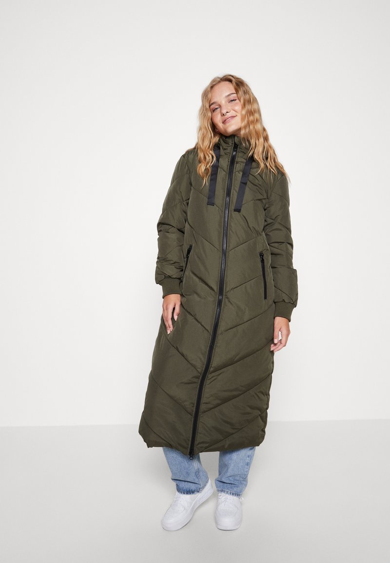 Casaco puffer longo verde oliva com costura em formato de chevron, capuz com cordão e fecho éclair preto. Usado com jeans azuis e sapatilhas brancas.