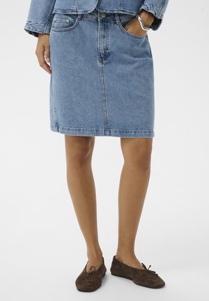 Žena v modrej rifľovej sukni po kolená, zodpovedajúcej rifľovej bunde, hnedých semišových balerínach, s jednou rukou v vrecku a strieborným náramkom na zápästí.