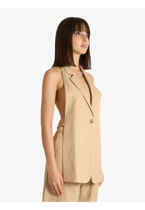 Giovane donna con capelli lisci e scuri indossa un gilet beige senza maniche con profondi tagli laterali e pantaloni beige coordinati, rivolta verso destra.
