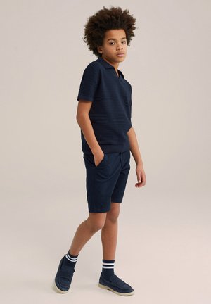 Polo shirt blu navy con un motivo testurizzato, abbinato a pantaloni corti blu navy. Il modello indossa mocassini blu navy e calze a strisce, con le mani in tasca.